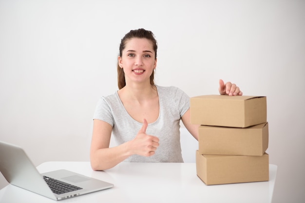 The girl on a light space sits at a table with a computer and corton boxes and shows thumb up. delivery service concept