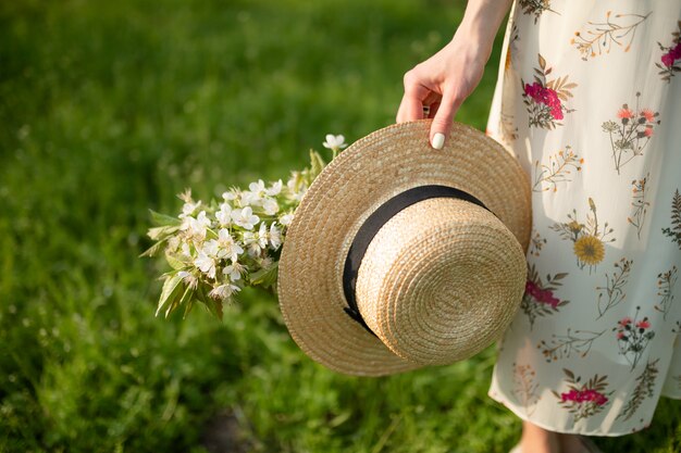 A girl in a light summer sundress walks in a spring green park enjoying the blooming nature, carries a wicker hat and flowers in her hand.