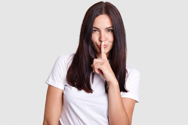 Photo girl like sly poses in photography. this photo is isolated. wall gray tint where model is in center. lady wears white t shirt that fits impeccably to manicure on her finger near mouth.