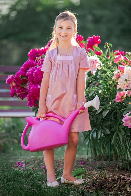 린넨 핑크 드레스를 입은 소녀는 정원에서 여름 맑은 저녁에 모란을 돌보고 꽃에 물을줍니다. 그는 아름답게 웃는다.