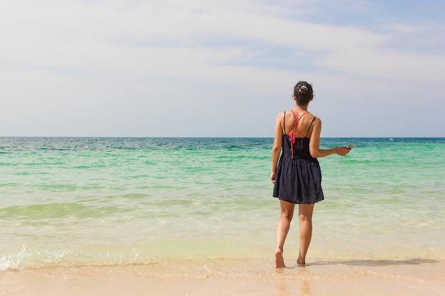 Girl listening to music on the beach facing the sea while holding mobile phone with earphones