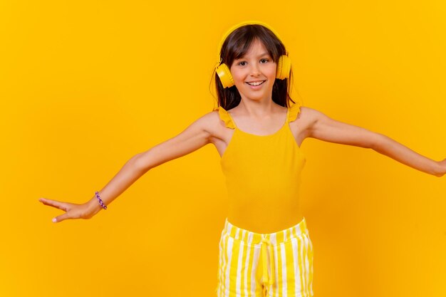 A girl listening to music and dancing with wireless headphones yellow background