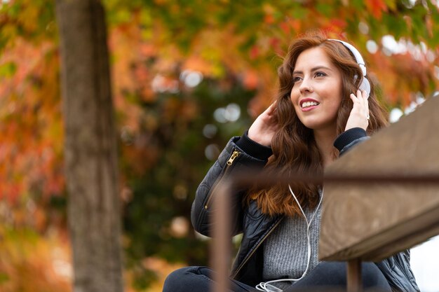 Girl listening to music with white headphones on a park bench in autumn with colorful trees