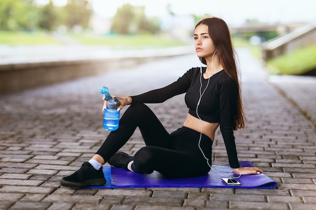 The girl listens to music while sitting on a gymnastic rug and rests after playing sports