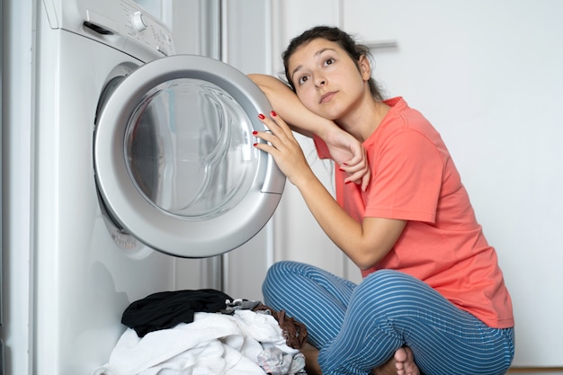 A girl loads dirty laundry into a washing machine while sitting\
on the floor in an apartment. laundry day, housework