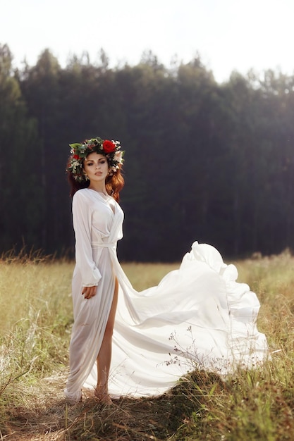 Girl in a long dress stands in a field with a wreath on her head and a bouquet of flowers in her han...