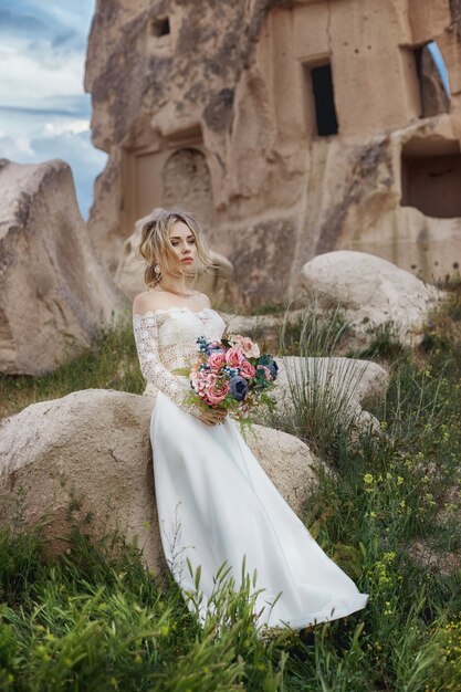 Girl in a long wedding dress with a bouquet of flowers in her hands sits in the mountains in nature.