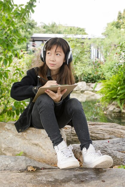 Girl looking away while writing over clipboard on rock against trees