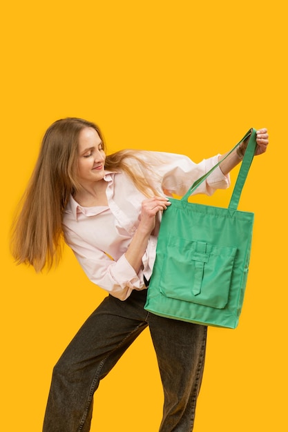 Девочка смотрит на сумку для покупок Молодая женщина с модной текстильной зеленой сумкой Студийный портрет, изолированный на желтом фоне