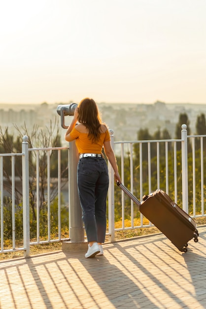 Girl looking through binoculars in park while holding her suitcase