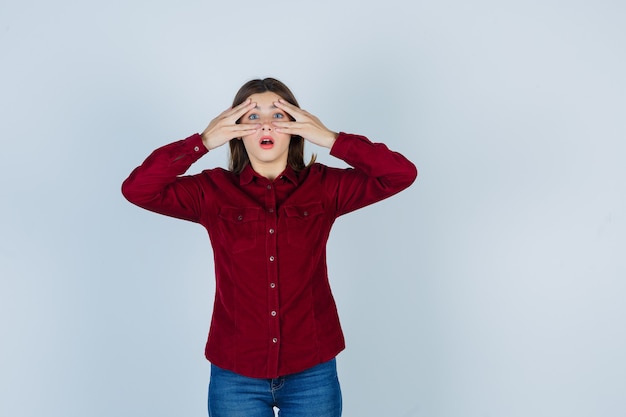 Girl looking through fingers in burgundy blouse and looking astonished.