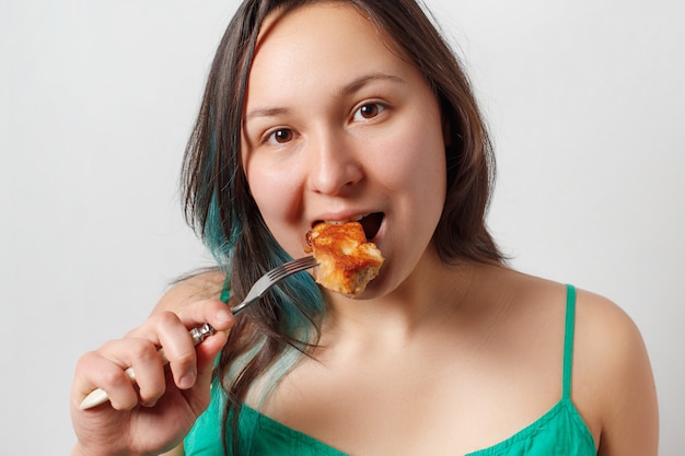 The girl looks in the frame and eats a piece of fried meat on a fork.