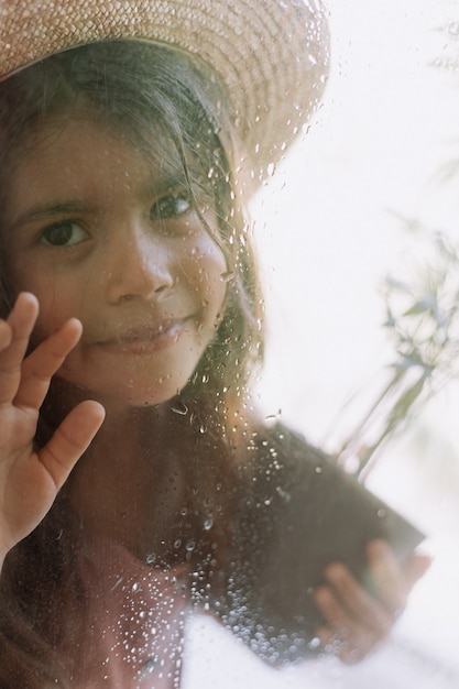 Photo girl looks out of the window from the house through the wet glass from the rain beautiful face