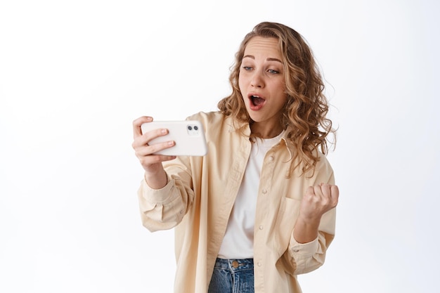 Girl looks at smartphone with amazement, watching video or stream, checking out awesome thing online, standing with mobile phone over white background