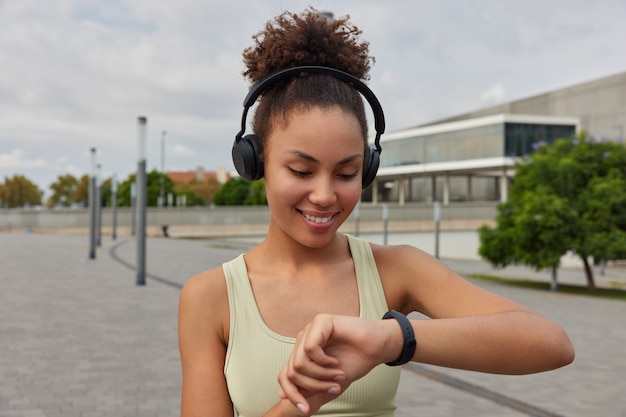 Photo girl looks at smartwatch after sport practice satisfied with results listens music during workout dressed in sportswear poses outdoors on stadium