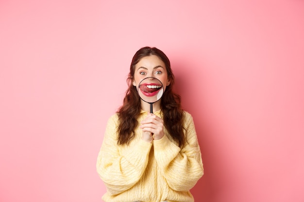 Girl looks at something tasty. Young woman shows her lips and tempted smile with magnifying glass, licking lip as staring at delicious food, standing against pink wall.