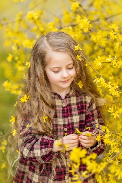 女の子は黄色い花を見ます。レンギョウを背景にした子供。彼女の髪に花を持つ子供の春の肖像画