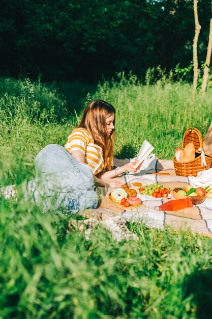 Девушка лежит у свежих фруктов, ягод, напитков и выпечки на одеяле для пикника на открытом воздухе