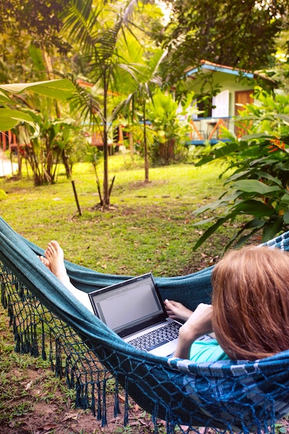 Ragazza sdraiata su un'amaca e lavorando su un computer portatile. vacanza