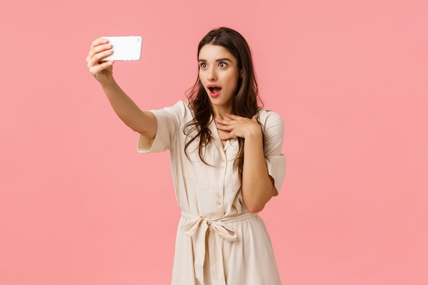 Ragazza che fa espressione affascinata e sorpresa come parlare con l'amica sulla videochiamata del telefono. affascinante donna bruna in abito, prendendo selfie, ansimando bocca aperta divertita, in piedi muro rosa