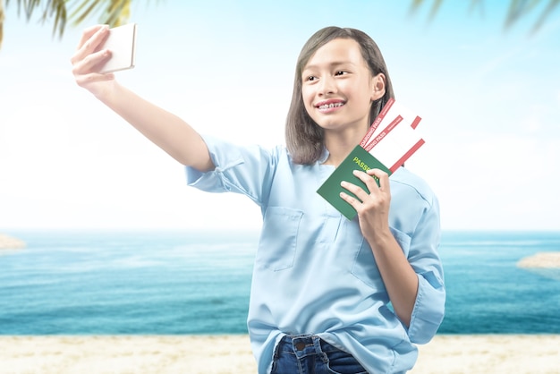 Girl making selfie holding ticket and passport on the beach with a blue sky background