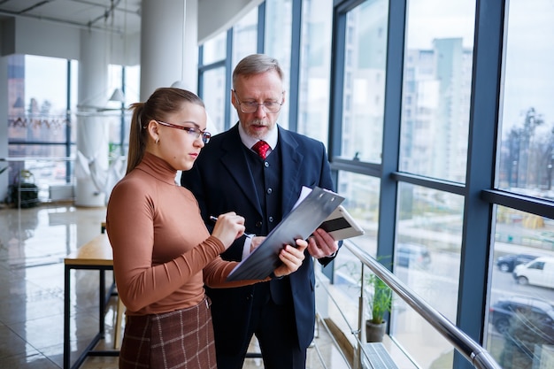 Girl manager in her own office tells a successful new business plan for economic development to her teacher mentor boss. Holds a folder with important documents in his hands.