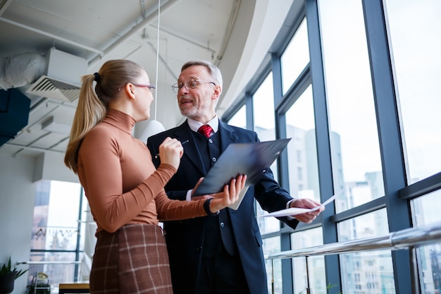 Girl manager in her own office tells a successful new business plan for economic development to her teacher mentor boss. Holds a folder with important documents in his hands.