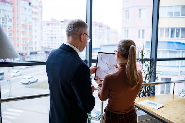 Girl manager in her own office tells a successful new business plan for economic development to her teacher mentor boss. Holds a folder with important documents in his hands.