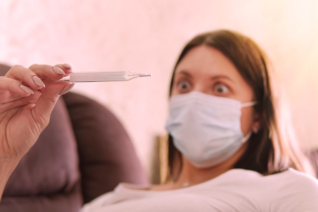 Ragazza in maschera da virus con termometro in mano. temperatura, termometro. la donna è sorpresa dall'alta temperatura sul termometro. paura di ammalarsi. emozione smarrimento. epidemia di coronavirus