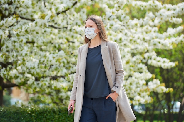 コロナウイルスの蔓延を防ぐために医療用マスクをした女の子が公園でスマートフォンを持っています。 COVID-19に対してフェイスマスクをした女性が、花の咲く木々の間の庭で社会的な距離を保ちます。