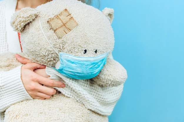 A girl in a medical mask holds a toy dressed in a medical mask on blue 