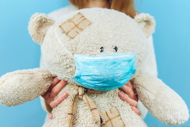 A girl in a medical mask holds a toy dressed in a medical mask on blue