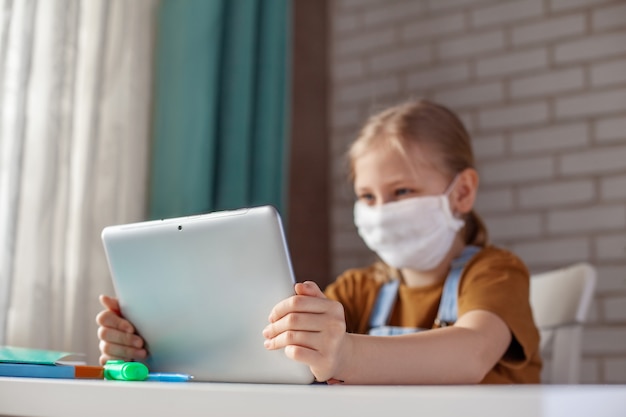 Una ragazza in una maschera medica sta studiando a casa con un laptop tavoletta digitale e sta facendo i compiti. formazione online a distanza