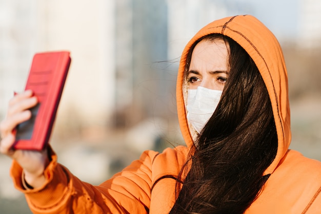 A girl in a medical mask photographs herself on the street during the pandemic of coronavirus and Covid - 19