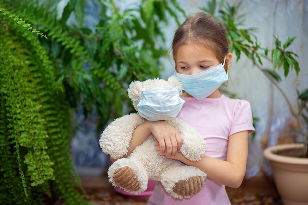 A girl in a medical mask sits at home during a Vieste epidemic with her teddy bear also in a mask