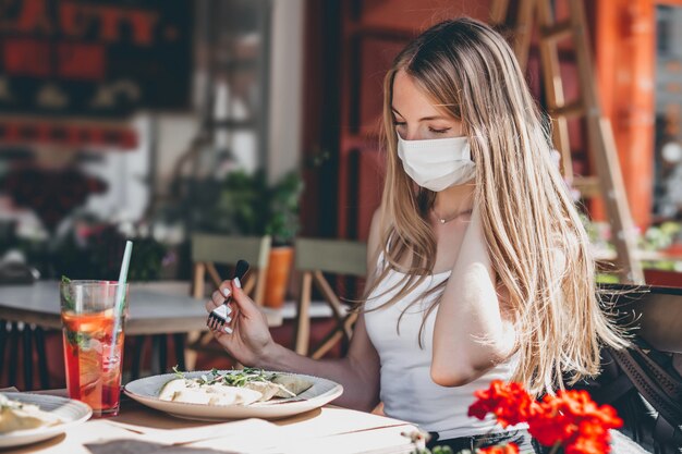 Girl in a medical mask sits at a table in a cafe and holds a fork and eats a dish on a plate