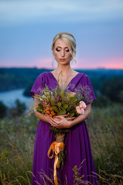 Girl model blonde in a lilac dress with a bouquet with a green forest