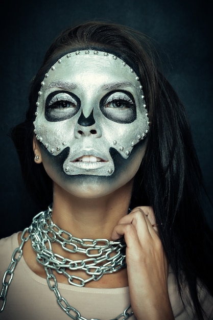 Girl model wearing chains with silver mask skull paint on dark background