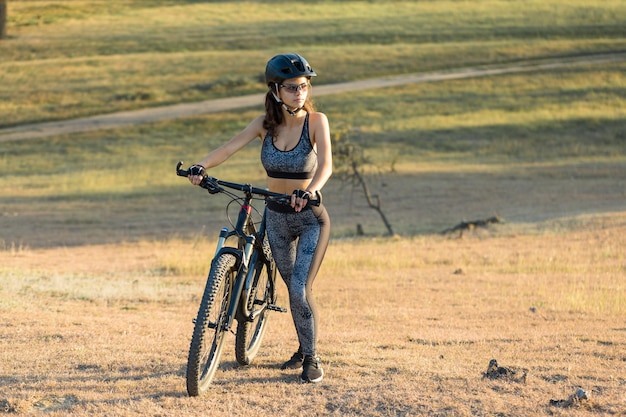 Girl on a mountain bike on offroad beautiful portrait of a\
cyclist at sunset fitness girl rides a modern carbon fiber mountain\
bike in sportswear closeup portrait of a girl in a helmet and\
glasses