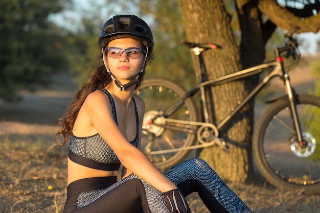 オフロードでマウンテンバイクに乗っている女の子日没時のサイクリストの美しい肖像画フィットネスの女の子がスポーツウェアでモダンなカーボンファイバーマウンテンバイクに乗る