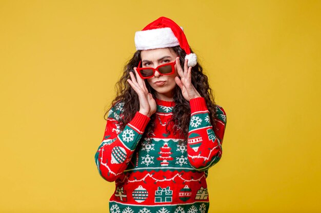 girl in New Year's clothes and santa hat puts on glasses on yellow background