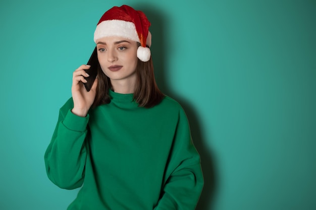 A girl in a New Year's hat is talking on the phone on a plain background holding a phone in her hand ordering dinner the concept of New Year's weekend 2023 happy christmas 2023 holidays