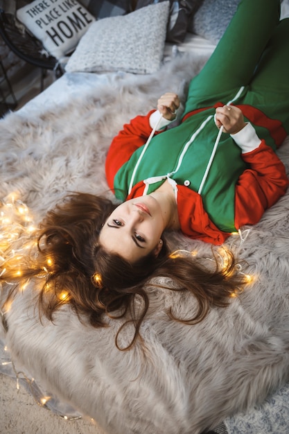 A girl in a New Year's overalls with a festive mood lies on a bed around luminous garlands