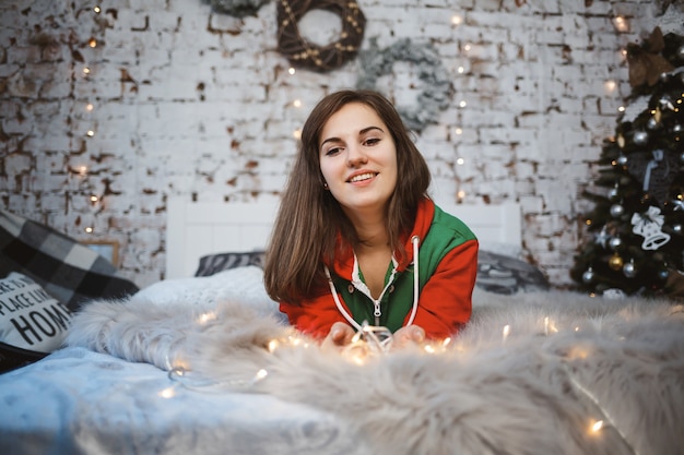 A girl in a New Year's overalls with a festive mood lies on a bed around luminous garlands