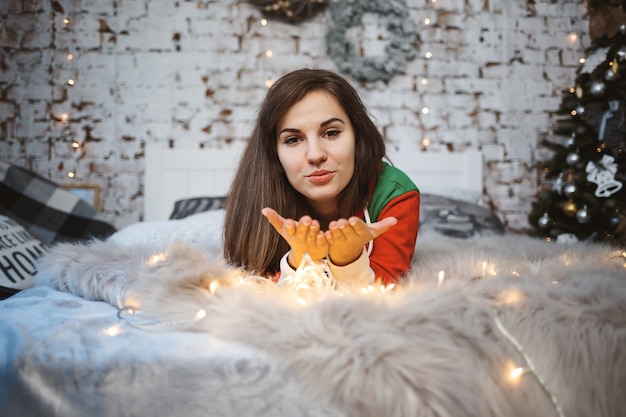 A girl in a New Year's overalls with a festive mood lies on a bed around luminous garlands