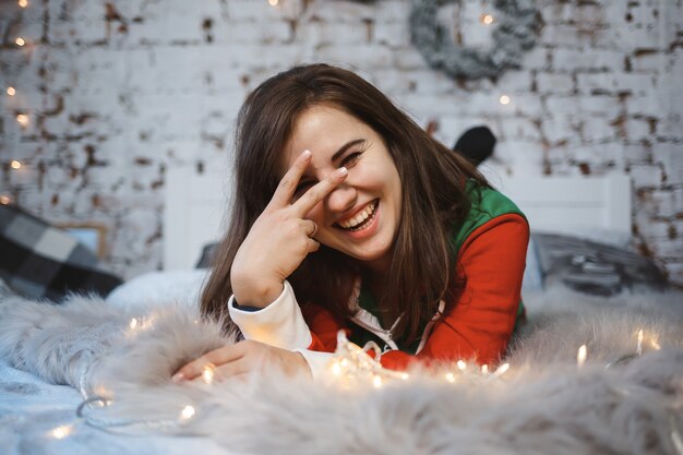 A girl in a New Year's overalls with a festive mood lies on a bed around luminous garlands