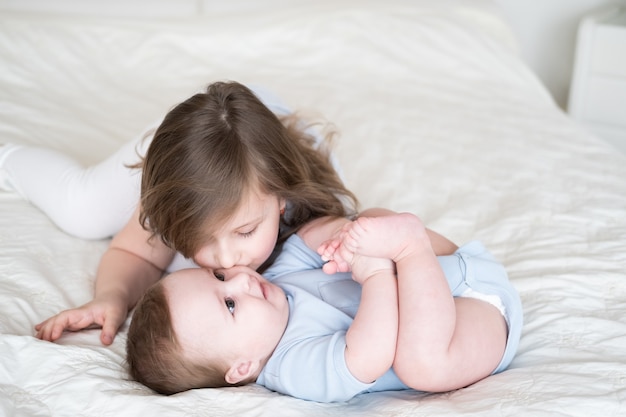 Girl older sister kissing her little baby boy brother on bed at home.