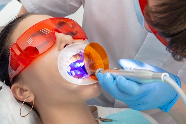 Девушка на осмотре у стоматолога. лечение кариозного зуба. врач пользуется зеркалом на ручке и борным аппаратом; брат-медик работает с полимеризационной лампой, чтобы укрепить пломбу