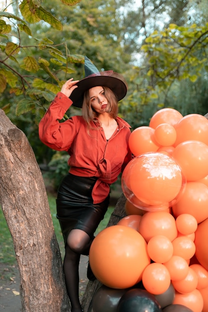 A girl in an orange shirt, a black skirt and a witch's hat on a background of balloons. Halloween