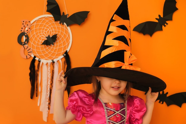 Photo a girl on an orange studio background puts on a witch hat for halloween hiding her face under it